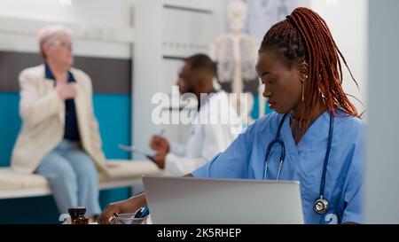 Infermiera afro-americana che lavora su appuntamenti in gabinetto, utilizzando il portatile per pianificare visite di controllo per aiutare i pazienti presso la struttura sanitaria. Assistente medico con consulenze di pianificazione stetoscopica. Foto Stock