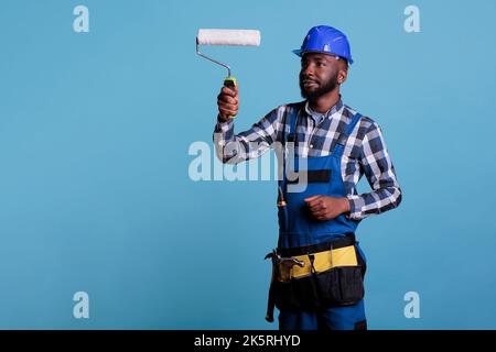 Il pittore si è concentrato sul lavoro che tiene il rullo professionale per dipingere una parete interna. Lavoratore di costruzione godendo il lavoro vestito in tute contro sfondo blu, ritratto studio. Foto Stock