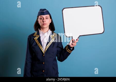 Sorridente stewardess che tiene la bolla bianca vuota di discorso, assistente di volo in piedi con la mock della cornice di comunicazione in su. Hostess aereo in uniforme aeronautica con messaggio vuoto palloncino, posto per il testo Foto Stock