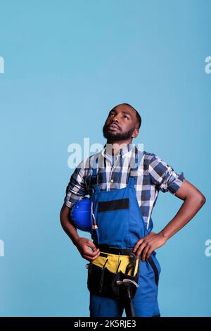 Lavoratore di costruzione attento che guarda verso l'alto tenendo il cappello indossare tute in studio girato su sfondo blu. Costruttore che pensa alle decisioni future prendendo una breve pausa dal lavoro. Foto Stock