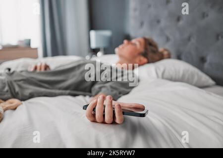 Telefono, letto e donna che dormono in una camera da letto, esausti e sofferenti con problemi di salute mentale. Stanco, pigro e procrastinato da donna depressa Foto Stock