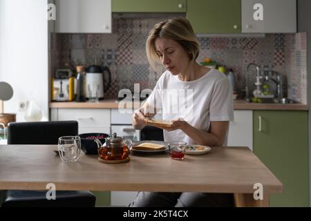 Donna di mezza età seduta al tavolo in cucina fa gustosa colazione con toast e tè in teiera Foto Stock