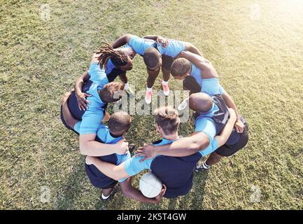Al di sopra di un gruppo diversificato di giocatori di rugby in piedi in un huddle insieme fuori su un campo. Giovani atleti maschi che guardano seri e concentrati mentre si sono accoccolati Foto Stock