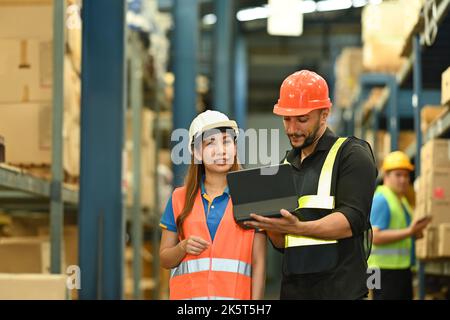 Dirigenti e lavoratori maschi che controllano le merci appena arrivate su un tablet digitale mentre si trovano vicino a scaffali alti in un grande magazzino Foto Stock