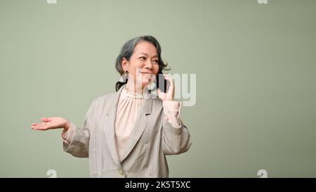 Splendida donna asiatica di 60s anni in tuta casual parlare al telefono con qualcuno, affari, contatto partner, in piedi isolato sul verde studio b Foto Stock