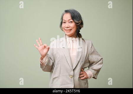 Splendida e bella 60s donna asiatica senior sorridente, mostrando OK segno mano e in piedi contro lo sfondo verde studio. Affare perfetto, Agreemen Foto Stock