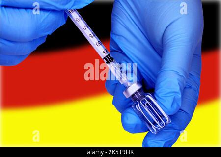 Mani ravvicinate in guanti medici blu prelevare un vaccino da un flaconcino in una siringa sullo sfondo della bandiera tedesca. Concetto di sciopero dei medici. Foto Stock