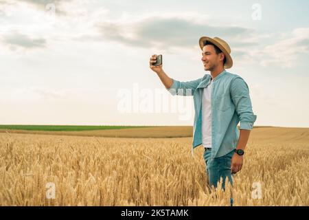 Felice agricoltore fotografando le colture con il telefono mentre si trova in piedi nel suo campo di grano crescente. Foto Stock