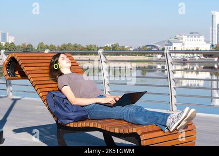 Una studentessa stanca si è addormentata su una panchina del parco mentre studiava online, guardando lezioni online su un computer portatile. La ragazza si addormenta per lavoro eccessivo. Onlin Foto Stock