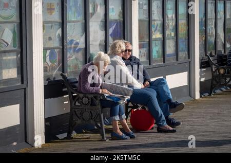 Una donna anziana siede su una panchina pubblica per leggere un giornale accanto a una coppia di mezza età a Worthing Pier, West Sussex, UK. Foto Stock