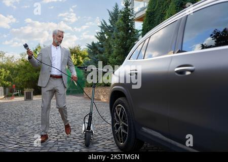 Uomo d'affari in tuta sul modo di lavorare con lo scooter elettrico e caricare la sua auto elettrica. Concetto di eco-pendolarismo e trasporto verde. Foto Stock