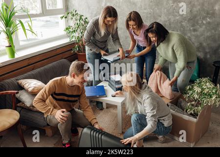 Amici sorridenti, energici, affollati che si accollano i bagagli e cercano di mettere le cose in valigia nel soggiorno, mostrare, entrare Foto Stock