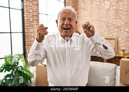 Uomo anziano con capelli grigi che tiene la moneta virtuale bitcoin urlare orgoglioso, celebrando la vittoria e il successo molto eccitato con le braccia sollevate Foto Stock