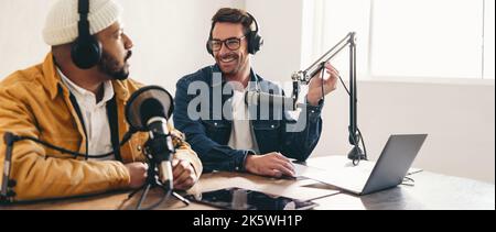 Buon presentatore della radio sorridente mentre intervistano un ospite su un podcast. Allegro giovane uomo co-hosting di una trasmissione audio con un ospite. Due crea di contenuti Foto Stock