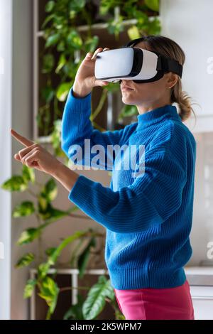 La giovane donna con coda di cavallo in maglione blu ama giocare a giochi interessanti in occhiali VR a casa Foto Stock