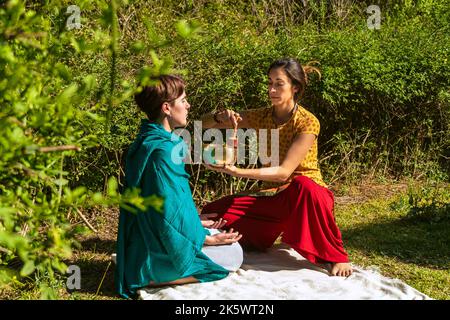 Terapeuta femminile concentrata in possesso di una ciotola di canto che esegue la terapia su una donna seduta in posizione lotus. Concetto di terapia di guarigione del suono. Foto Stock