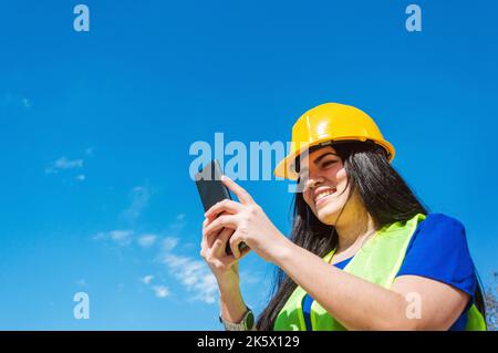 giovane lavoratrice latina, caucasica, venezuelana con elmetto di sicurezza giallo all'aperto felice sorridendo controllando il suo telefono, leggendo le notifiche, con blu Foto Stock