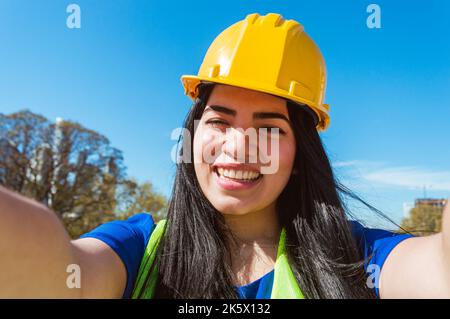 ritratto di una giovane donna operaia caucasica venezuelana, con abiti blu, casco giallo e giubbotto di sicurezza è all'aperto sorridendo prendendo un selfie con lui Foto Stock