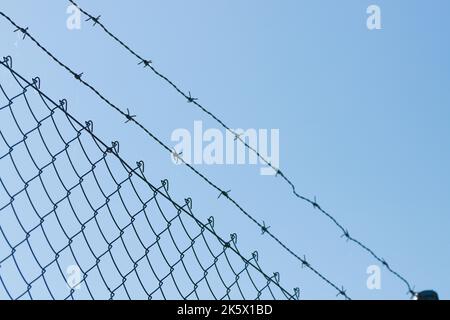 Vista diagonale di una recinzione realizzata con rete metallica e filo spinato in cima, isolata su sfondo blu cielo Foto Stock