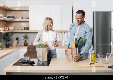 Ritratto di felice giovane coppia sposata che viene a casa da shopping e disimballaggio sacchetti di carta con alimentari in cucina insieme. Concetto di famiglia e di rapporto. Foto Stock