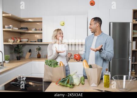 Giovane arabo e donna in abbigliamento casual cucina insieme a casa da prodotti sani e freschi. Buona famiglia che prepara insalata fresca, pasto in cucina moderna. Arabo che lancia i pomodori per divertimento. Foto Stock