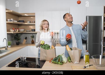 Giovane arabo e donna in abbigliamento casual cucina insieme a casa da prodotti sani e freschi. Buona famiglia che prepara insalata fresca, pasto in cucina moderna. Arabo che lancia i pomodori per divertimento. Foto Stock