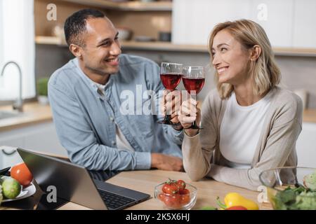 Una cena di cucina per tutta la famiglia in una casa moderna e luminosa. Coppia innamorata, donna caucasica bionda e bel sorriso arabo che parla e fare insalata, bere vino rosso e godere di una giornata perfetta. Foto Stock