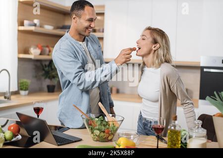 Coppia multinazionale sposata allegra usando il laptop mentre cucinava cibo sano in cucina, bei sposi giovani sposi che si divertono mentre nutrono sua moglie con pomodoro ciliegia, preparando pasto vegetale Foto Stock