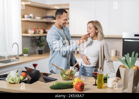 Coppia multinazionale sposata allegra usando il laptop mentre cucinava cibo sano in cucina, bei sposi giovani sposi che si divertono mentre nutrono sua moglie con pomodoro ciliegia, preparando pasto vegetale Foto Stock