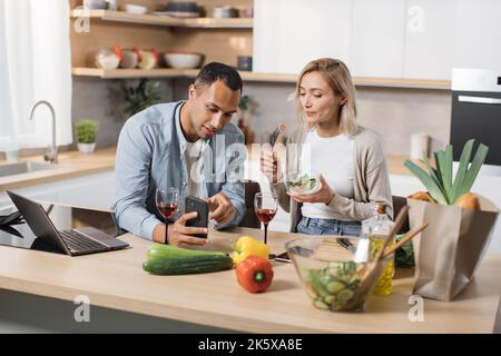 Giovane coppia attraente innamorata mangiare insalata da verdure fresche utilizzando lo smartphone mentre si effettua una videochiamata online con parenti o amici in cucina moderna a casa. Foto Stock