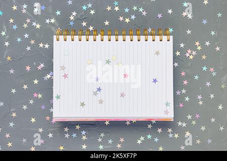 Apri la pagina vuota del blocco note su sfondo nero scuro festivo con stelle colorate. Per fare l'elenco, la scrittura e il concetto di narrazione, copia lo spazio Foto Stock