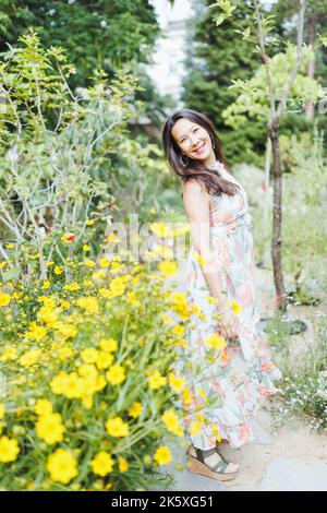 Bella felice gara mista donna giapponese in romantico abito floreale maxi e su zeppa tacchi in giardino estivo, sorridente. Ripresa a tutto corpo. Luce del sole, spazio di copia Foto Stock