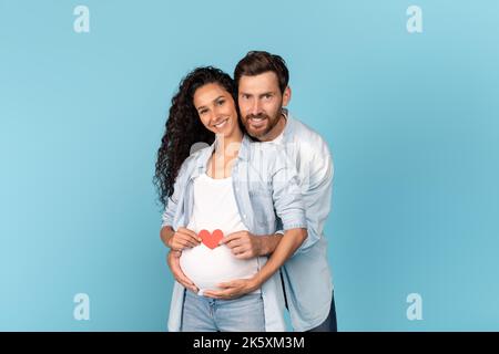 Felice uomo caucasico millenario con barba che abbraccia una donna arabica incinta, tenendo il cuore sul ventre Foto Stock