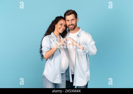 Giovane ragazzo caucasico felice con la barba abbraccia moglie araba incinta, fare le mani segno in forma di cuore Foto Stock