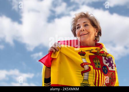 Donna anziana sorridente, circondata dalla bandiera di Spagna su uno sfondo blu con nuvole. Orgoglio di essere spagnolo. Amore e patriottismo. Columbus giorno Foto Stock