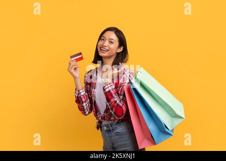 Concetto di rimborso. Donna asiatica felice posa con carta di credito e borse shopping colorate su sfondo giallo studio Foto Stock