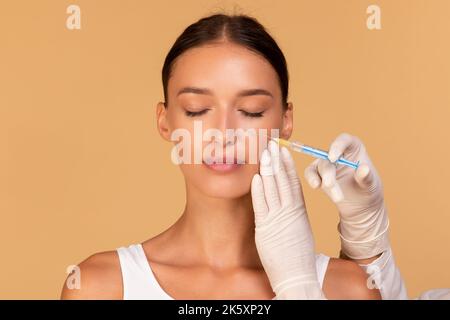 Chirurgia plastica e concetto di iniezione di bellezza. Attraente giovane donna che riceve botox per le rughe, sfondo beige Foto Stock