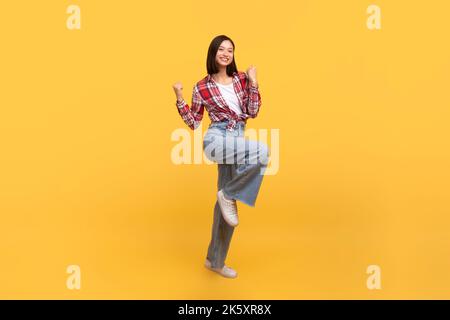 Donna asiatica entusiasta celebrare il successo e saltare in aria, felice di divertirsi su sfondo giallo studio, a tutta lunghezza Foto Stock