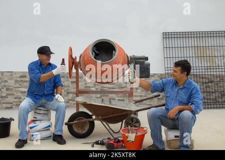 Due operai edili, uno giovane e uno anziano durante la pausa pranzo mentre brinda con una birra. Sorridere i lavoratori di un cantiere di costruzione surrogato Foto Stock