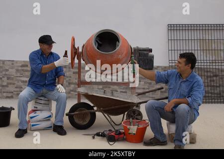 Due operai edili, uno giovane e uno anziano durante la pausa pranzo mentre brinda con una birra. Sorridere i lavoratori di un cantiere di costruzione surrogato Foto Stock