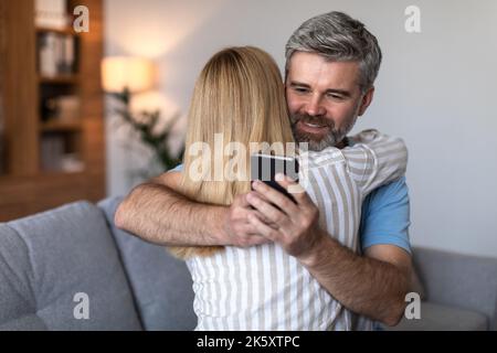 Allegro uomo europeo di mezza età che abbraccia una donna e digita sullo smartphone nel salotto interno Foto Stock