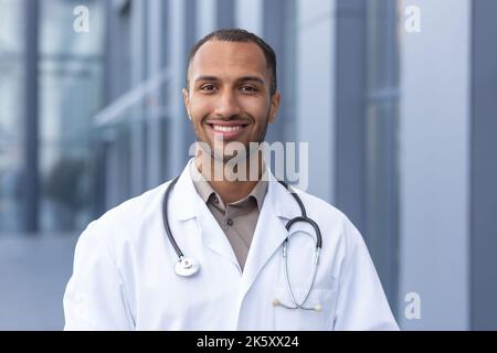 Ritratto di un medico afro-americano riuscito e sorridente, uomo in cappotto medico con stetoscopio che guarda la macchina fotografica e sorridente, al di fuori del moderno ospedale. Foto Stock