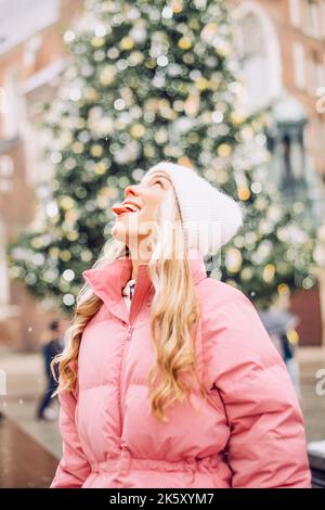 Un carino ritratto di una ragazza in inverno sulla strada, la neve sta cadendo, la bionda sullo sfondo dell'albero di Natale cattura fiocchi di neve con Foto Stock