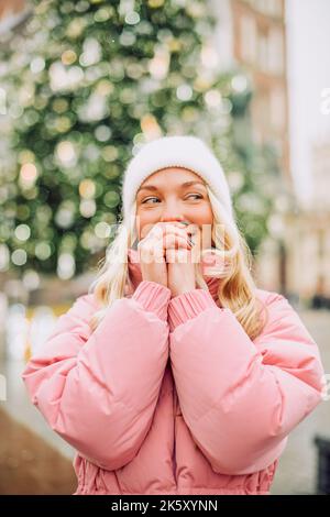 Carino ritratto di una ragazza in inverno sulla strada, la neve sta cadendo, la bionda, albero di Natale, riscalda le mani. Ragazza in un cappello bianco, jacke rosa Foto Stock