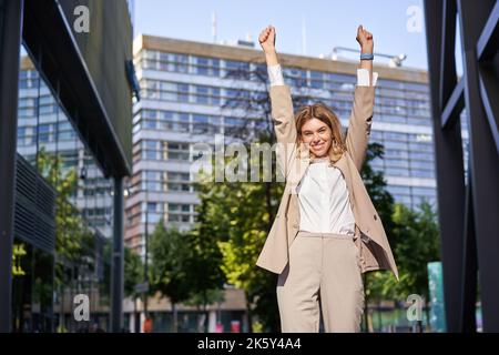 Ritratto di felice giovane donna d'affari alzare le mani, pugno pompa, celebra la vittoria, vince sth, raggiunge obiettivo, si trova all'aperto sulla strada Foto Stock