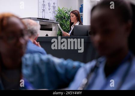 Receptionist asiatico utilizzando documenti di rapporto medico per aiutare i pazienti con i moduli di registrazione e visite di controllo. Lavoro al banco della reception dell'ospedale con stampante per fotocopie e computer per appuntamenti. Foto Stock