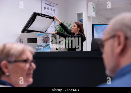 Il segretario utilizza una stampante presso il banco della reception per redigere i documenti prima delle consultazioni di controllo. Donna addetta al ricevimento che lavora su appuntamenti sanitari con moduli presso la reception. Foto Stock