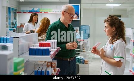 Farmacista femminile aiutare il gruppo multietnico di clienti a comprare farmaci e vitamine in farmacia, parlando di pillole sugli scaffali. Fornire assistenza con prodotti farmaceutici e farmaci. Foto Stock