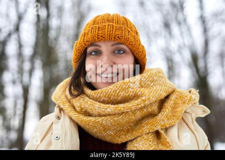 Ritratto di una giovane donna caucasica che indossa un cappello a maglia e una sciarpa nella foresta invernale e sorride alla macchina fotografica Foto Stock