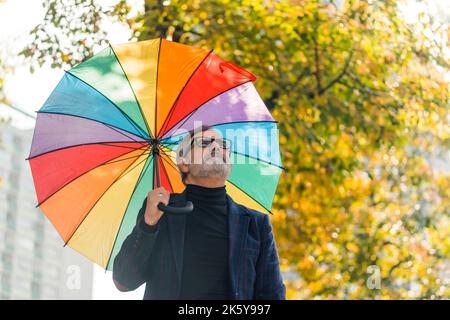 Tempo di autunno volatile. Pigro Domenica a piedi nel vicino parco. Foto da esterno di un elegante uomo caucasico bearded con occhiali neri che tengono un grande ombrello arcobaleno vivace e che guarda il cielo. Sfondo sfocato con albero che ha cambiato il colore delle foglie. Foto di alta qualità Foto Stock